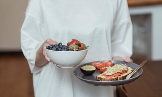 Person holding a bowl of fresh berries and a plate with avocado toast, displaying a healthy breakfast spread.